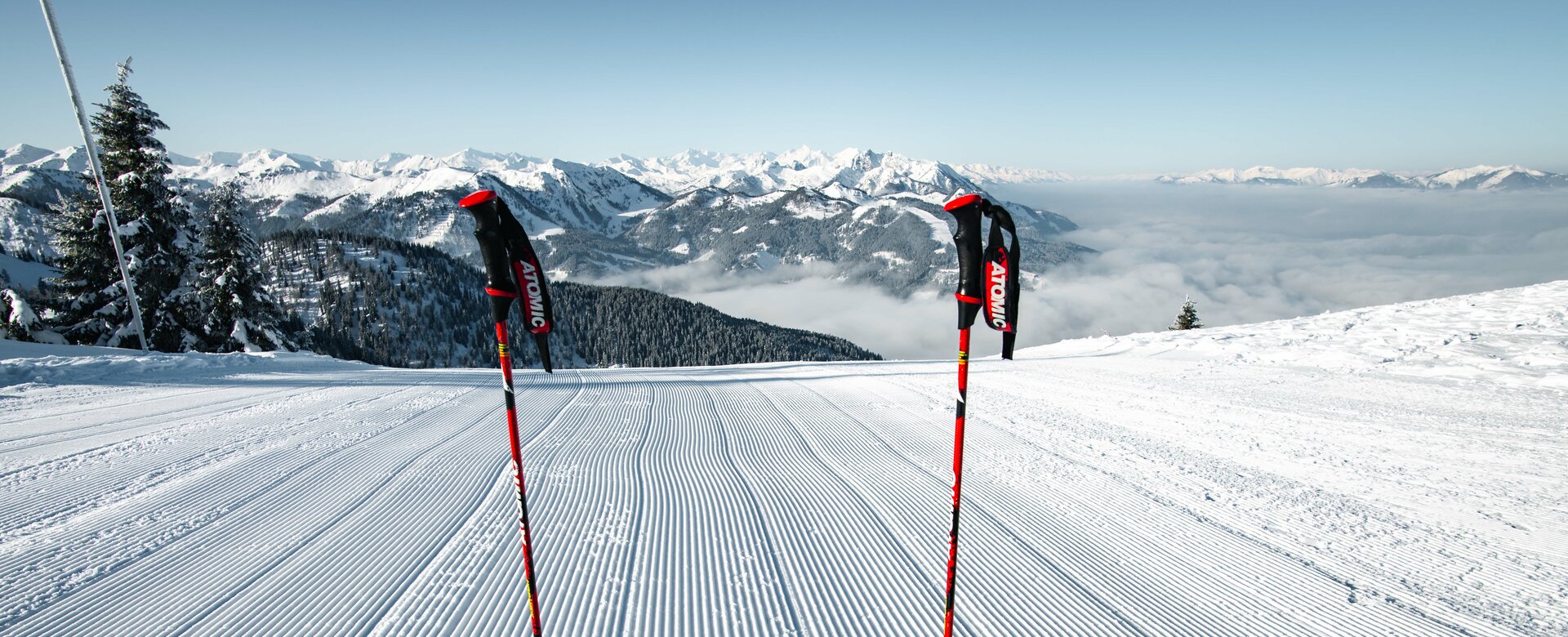 Two ski poles are stuck in the freshly groomed piste and the ski tip can also be seen as well as a snow-covered mountain landscape in the distance | © Snow Space Salzburg Christian Schartner
